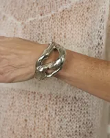 Brazalete abierto de diseño orgánico y voluminoso, con acabado metálico brillante. Su forma irregular y ergonómica permite un ajuste cómodo en la muñeca o antebrazo.