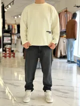 Sweater de punto liviano color beige, con textura marcada, cuello redondo y mangas largas de corte relajado, con pequeño estampado tipográfico negro en el lateral inferior.