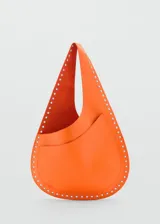 Bolso de hombro tipo hobo en color naranja, confeccionado en piel bovina. Presenta un diseño de silueta redondeada con detalle de tachuelas metálicas plateadas en todo el borde y un bolsillo exterior abierto.