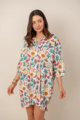 Vestido corto blanco con diseño estampado de temática tropical y mexicana que incluye loros, cactus, palmeras, guitarras y sombreros. Presenta cuello camisero, cierre frontal con botones y mangas tres cuartos de corte holgado.