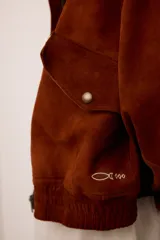 Chaqueta de gamuza color marrón con cierre frontal, cuello alto y puños elásticos. Presenta un diseño de espíritu artesanal con apliques bordados en los bolsillos delanteros y en la espalda, y se encuentra completamente forrada.