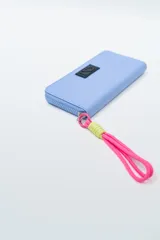 Billetera rectangular con cierre, color celeste pálido, con parche frontal negro que lleva las letras "CNJ" en relieve. Incluye un llavero de muñequera con cordón trenzado en rosa fuerte y verde lima.