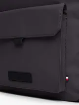 Mochila Tommy Hilfiger de diseño minimalista y silueta tipo domo en color gris oscuro. Cuenta con compartimento principal con cierre de cremallera, un bolsillo exterior frontal con solapa y cierre magnético, correas de hombro acolchadas y ajustables, y un asa superior. Presenta el logo de la marca en relieve en el frente y detalles distintivos.