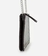 Cartera bandolera Satinato de color marrón oscuro, confeccionada en poliuretano con textura geométrica símil cocodrilo y detalles de tachas metálicas plateadas en los bordes. Cuenta con cierre superior y correa larga de cadena metálica.