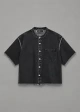 Chaqueta de denim negro con lavado ácido, cuello mao, mangas cortas, cierre frontal con botones metálicos y un bolsillo aplicado en el pecho. Presenta detalles de terminaciones desflecadas en el cuello, mangas y ruedo.
