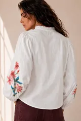 Camisa blanca de manga larga con bordados florales en tono rojo y verde en el frente y las mangas. Presenta cuello mao con escote en V y botones frontales.