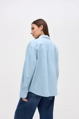 Camisa de algodón en color celeste claro, con cuello solapa, cierre frontal de botones y bordado pequeño en el pecho. Presenta un corte holgado y mangas largas.