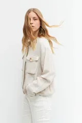 Chaqueta bomber color beige melange, de lino, con cuello mao, cierre frontal con botones a presión, dos bolsillos con solapa en el pecho y puños y cintura elastizados.