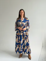 Vestido largo azul con estampado de hojas en tonos beige y blanco, escote en V con botones frontales, mangas largas amplias y diseño escalonado.