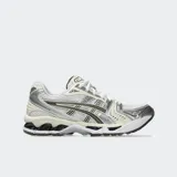 Championes de running Asics modelo Gel Kayano 14, con diseño retro inspirado en los años 2000. Presenta una base de malla blanca con superposiciones en tonos plateados y detalles en negro. Incorpora tecnología GEL en la suela para una amortiguación superior.