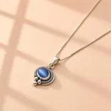 Collar de cadena fina en plata 925 con un dije circular de estilo étnico que presenta una piedra central azul con efecto ojo de gato, rodeada por un borde de filigrana y detalles de pequeñas esferas plateadas en la parte inferior.
