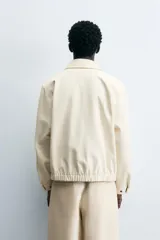 Cazadora de hombre color beige claro, corte relajado, con cuello solapa, manga larga y puños con botones a presión. Presenta bolsillos de plastrón con solapa en la cadera y cierre frontal de cremallera oculta por solapa de botones a presión.