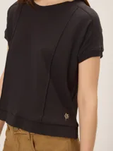 Remera tejida color beige de corte relajado, con mangas cortas y detalle de costuras frontales marcadas. Incluye un pequeño bordado distintivo en el borde inferior.