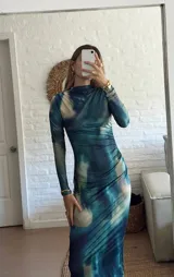 Vestido midi de manga larga con cuello alto, confeccionado en tela elastizada con estampado tipo tie-dye en tonos azules y beige. Presenta un diseño ajustado al cuerpo con frunces laterales en la zona de la cintura y cadera.