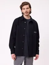 Camisa de gabardina color verde oliva, con cuello clásico, cierre frontal de botones y dos bolsillos frontales con parche. Incluye un pequeño parche con el logo de la marca en el bolsillo izquierdo.