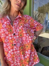 Camisa unisex de manga corta con cuello, confeccionada en lino y algodón con estampado floral en tonos naranja, rosa y blanco. Tiene botones de madera y corte holgado.