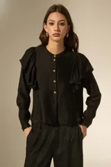 Camisa negra de viscosa con textura arrugada, cuello a la base y cierre frontal con botones. Presenta volados en los hombros y mangas largas.