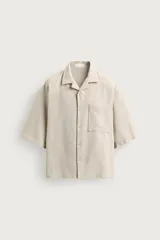 Camisa boxy fit de color beige, confeccionada en tejido con lino, con cuello bowling, mangas cortas, un bolsillo aplicado en el pecho y cierre frontal con botones.