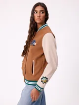 Campera estilo varsity con cuerpo en color marrón y mangas en tono beige. Presenta cierre frontal con botones a presión, cuello, puños y ruedo con franjas tejidas en verde y blanco. Incluye un parche con la letra R en el pecho y detalles bordados en las mangas.