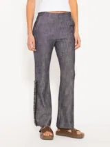 Pantalón de lino con apariencia denim, tiro bajo y corte recto. Presenta detalles de galón con flecos y cadenas en los laterales inferiores.