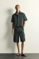 Bermuda cargo relaxed fit confeccionada en denim de mezcla de algodón y lino, con diseño de rayas verticales grises y negras. Cuenta con bolsillos laterales tipo cargo con solapa y cierre frontal con cremallera y botón.