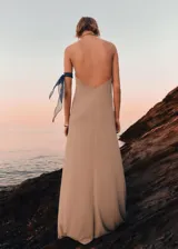 Vestido largo de diseño evasé en color beige, con cuello halter cruzado y espalda abierta. Presenta cierre de botones en el cuello.