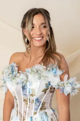 Vestido largo de fiesta en gasa de seda natural color blanco con estampado de flores celestes y verdes. Corsé strapless con ballenas y detalle de flores en escote y mangas caídas. Espalda cruzada y falda amplia con tajo delantero.