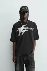 Camiseta negra de corte relaxed fit, cuello redondo y manga corta. Presenta un estampado frontal en blanco con efecto spray que simula dos figuras volando, y texto en un óvalo que dice "SUCCESS CHASER".