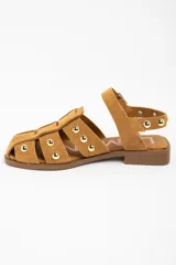 Sandalia tipo cangrejera de gamuza color camel, con diseño de tiras entrelazadas y tachas metálicas doradas. Cuenta con cierre de pulsera con hebilla ajustable y suela plana de goma.