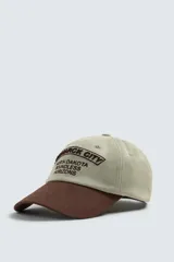 Gorra de seis paneles de algodón en color beige con visera marrón contrastante. Cuenta con un diseño bordado en marrón en el frente con texto alusivo.