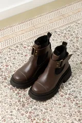 Bota tipo Chelsea de cuero marrón con suela track negra de goma. Presenta paneles elásticos laterales y una costura reforzada en la puntera. Incluye una correa de cuero desmontable con hebilla metálica.