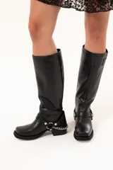 Bota estilo biker de cuero negro con caña alta y cierre metálico lateral. Presenta un diseño versátil con estribo y cadena desmontables, taco cuadrado y suela con plataforma.