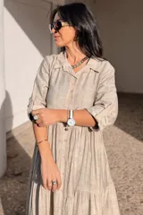 Vestido largo de estilo camisero en color azul, con cuello clásico, cierre frontal de botones y mangas tres cuartos con trabillas. Presenta un diseño de falda con cortes horizontales que le otorgan volumen y movimiento.