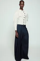 Pantalón de tiro medio de corte wide leg, confeccionado en mezcla de lyocell con patrón de rayas diplomáticas. Presenta bolsillos laterales ocultos, pliegues delanteros marcados y cierre frontal con cremallera, botón y ganchos metálicos.