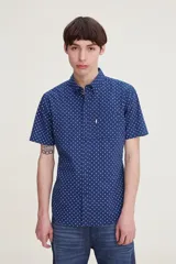 Camisa de manga corta color azul con estampado de pequeños rombos blancos. Presenta cuello clásico abotonado, cierre frontal con botones y un bolsillo en el pecho.