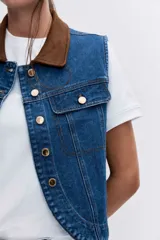 Chaleco de denim azul con cuello camisero en contraste color marrón, cierre frontal con botones metálicos y bolsillos superiores con solapa.