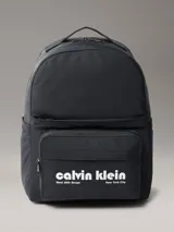 Mochila negra de poliéster con diseño moderno y funcional. Presenta un compartimento principal con cierre, bolsillos exteriores con cremallera en el frente, laterales y espalda, y correas acolchadas. En el bolsillo frontal se destaca el logo de Calvin Klein en blanco con la inscripción "West 39th Street New York City".