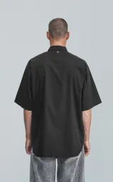 Camisa de manga corta en popelina de algodón, con diseño oversize y cierre frontal de botones. Presenta dos bolsillos delanteros, uno con solapa y otro con ojal y botón.