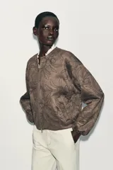 Chaqueta bomber color marrón con cuello redondo, manga larga con puño elástico y cierre frontal de cremallera metálica. Presenta un diseño de bordados florales en relieve por toda la prenda y bolsillos delanteros de vivo.