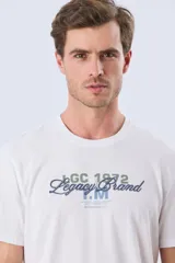 Remera azul marino de manga corta con cuello redondo, confeccionada en mezcla de algodón y poliéster. Presenta un diseño en el frente que combina estampa y bordado con la leyenda "Legacy Brand 1972" en tonos rosado, verde y marrón.