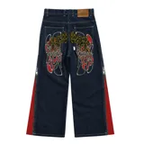 Pantalón de jean azul raw con diseño de corte versátil, ajustable mediante cierres laterales que revelan un panel interior rojo. Presenta una estampa gráfica de gran tamaño en la parte trasera con detalles bordados, costuras reforzadas y etiqueta de cuero en la cintura.
