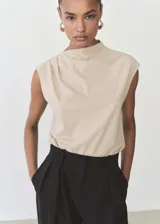 Camiseta color beige con cuello alto asimétrico, sin mangas y detalles fruncidos. Tejido fluido y diseño recto.