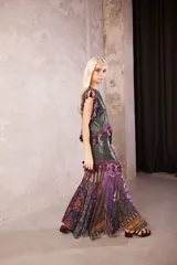 Falda larga de cuero verde oliva con diseño de paneles. La parte superior es de cuero liso con cierre frontal, decorada con un galón de borlas en tonos morados y verdes. La parte inferior presenta una falda de tela fluida con estampado floral y geométrico en tonos violeta, morado y verde.