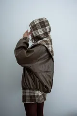 Chaqueta de cuero sintético color marrón con capucha y solapas amplias en tela escocesa a cuadros. Posee cierre frontal, puños elásticos y diseño de corte holgado.