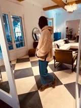 Sweater beige de corderito con capucha y bolsillo frontal de cuero en tonos marrón y ocre. Presenta tajos laterales y cierre parcial con cremallera en el cuello.