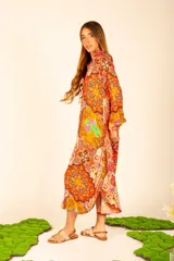 Vestido camisero midi color naranja con estampado floral y paisley. Tiene cuello camisero, mangas largas con puños abotonados, bolsillos laterales y aberturas laterales en el ruedo.