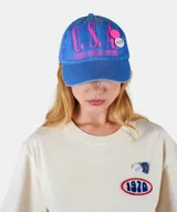 Gorra de algodón azul con efecto desgastado, con inscripción frontal 'U.S.A.' en color fucsia y apliques decorativos tipo pins. Cuenta con visera curva y cierre ajustable.