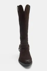 Bota alta de serraje marrón oscuro, de estilo texano, con tacón en bloque y puntera ligeramente puntiaguda. Cuenta con detalle de tiras cruzadas con tachas metálicas y una arandela en el tobillo, además de trabillas laterales en la parte superior de la caña.