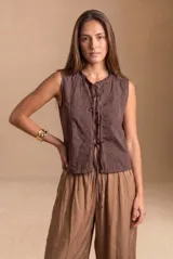 Blusa de algodón sin mangas en color blanco off-white, con cuello alto con volados y cierre frontal de cordones ajustables con lazos. Presenta detalles de encaje y bordados sutiles en la parte delantera.