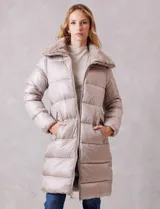 Campera puffer larga de acabado satinado en tono beige. Presenta un diseño capitoneado horizontal, cierre frontal bidireccional y un amplio cuello envolvente revestido en piel sintética. Incluye bolsillos laterales funcionales con cierre y logo metálico en la manga.